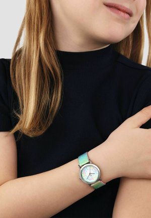 Child wearing a multicolored gradient watch with large, colorful numbers and a black short-sleeve top, arms crossed gently.