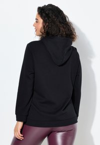 Donna con capelli ricci che indossa una felpa nera e leggings viola lucidi, di profilo su uno sfondo bianco.