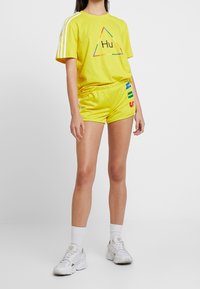 Gul t-shirt med triangul "Hu" grafik, i kombination med matchande shorts med färgglad text längs sidan. Vita sneakers fulländar looken.