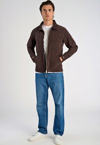 Chaqueta de pana marrón con cuello, bolsillo delantero y puños de botón, llevada sobre una camisa blanca y jeans azules con zapatillas blancas.