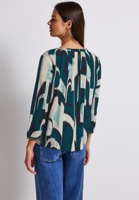 Blouse imprimée avec des nuances de bleu sarcelle, crème et marine. Tissu léger avec une coupe décontractée et des manches trois-quarts. Détail de col froncé.