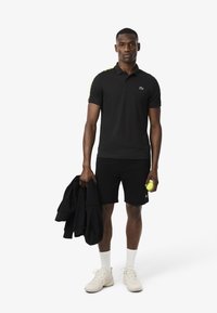 Polo shirt nera con dettagli verdi, abbinata a pantaloni corti neri. Calze bianche e scarpe da ginnastica chiare. Tiene in mano una giacca nera e una pallina da tennis gialla.