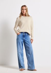 Beżowy sweter dzianinowy z okrągłym dekoltem, zestawiony z luźnymi jasnoniebieskimi dżinsami. Posiada szerokie nogawki i boczne kieszenie. Ubrane z płaskimi sandałami.