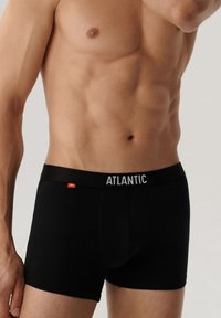 Męski tors w czarnych bokserkach z logo "ATLANTIC" na pasku, na jednolitym jasnym tle.