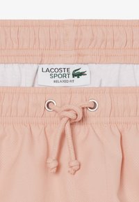 Hellrosa Lacoste Sport Shorts mit elastischem Bund, Kordelzug, der zu einem Knoten gebunden ist, und sichtbarem Etikett mit der Aufschrift "Relaxed Fit" sowie dem Krokodil-Logo.