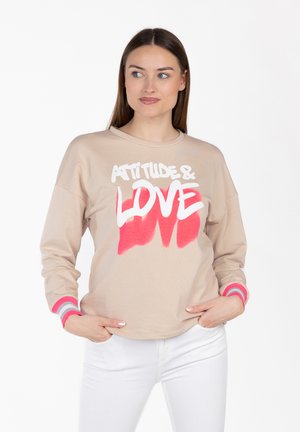 Vrouw draagt beige sweatshirt met de tekst "Attitude & Love" in wit en roze, gecombineerd met een witte broek, handen in de zakken, kijkt opzij.