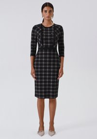 Zwarte geruite jurk met een getailleerd silhouet, driekwart mouwen en decoratieve knopen aan de voorkant; gecombineerd met lichtgekleurde puntige hakken.