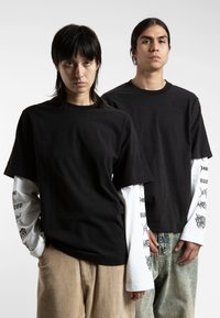Zwarte oversized katoenen t-shirt gelaagd over een witte lange mouwen met bedrukte accenten. Het model draagt een beige corduroy broek, die een ontspannen pasvorm toont.