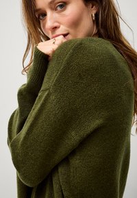 Maglione a maglia verde oliva con una texture morbida e un fit rilassato, caratterizzato da spalle scese e maniche lunghe, indossato aderente al corpo.