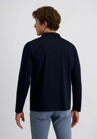 Mann mit welligem braunem Haar, der ein lang�ärmliges navyblaues Shirt und hellblaue Jeans trägt, steht mit dem Rücken zu einer schlichten weißen Wand.