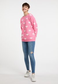 Maglione rosa con motivi a cuore bianchi, colletto tondo e polsini a coste, abbinato a jeans skinny blu strappati e sneakers bianche.