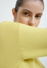 Sweter robiony na drutach w pastelowym żółtym kolorze, z teksturowanym wzorem. Zbliżenie na rękaw i ramię, podkreślające jego luźny krój i miękki materiał.