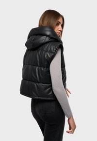 Gilet imbottito nero con cappuccio, realizzato in materiale lucido e trapuntato. Design senza maniche con dettagli di cucitura prominenti. Indossato sopra una maglietta a maniche lunghe grigia.