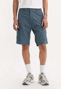 Osoba v modrých cargo šortkách, bílých ponožkách a šedobílých sportovních botách, stojící s uvolněnýma rukama podél těla a v bílém tričku.