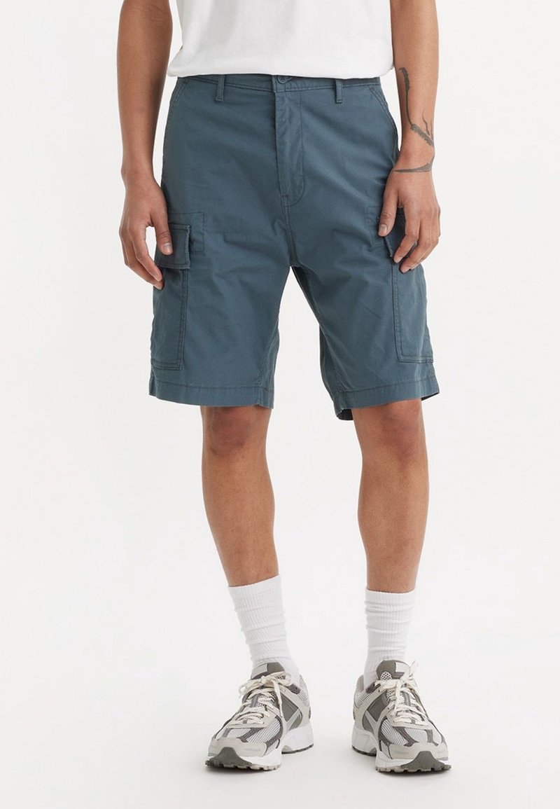 Osoba v modrých cargo šortkách, bílých ponožkách a šedobílých sportovních botách, stojící s uvolněnýma rukama podél těla a v bílém tričku.