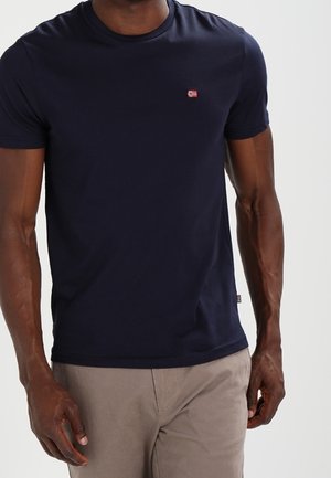 Mann trägt ein marineblaues T-Shirt mit kurzen Ärmeln mit kleinem rot-weißem Logo auf der linken Brust und beigefarbene Hose.