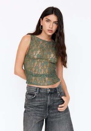 Top en dentelle verte avec une silhouette ajustée, un col rond et une texture superposée, associé à un jean en denim foncé à taille haute.