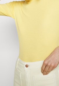 Gul ribbet turtleneck-top parret med højtaljede cremede bukser, der har en metalknap i taljen og sidelommer.