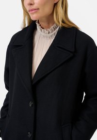 Cappotto nero texture con colletto a risvoltino, chiusura frontale con bottoni e tasche laterali, indossato sopra una blusa beige a collo alto con pieghe.