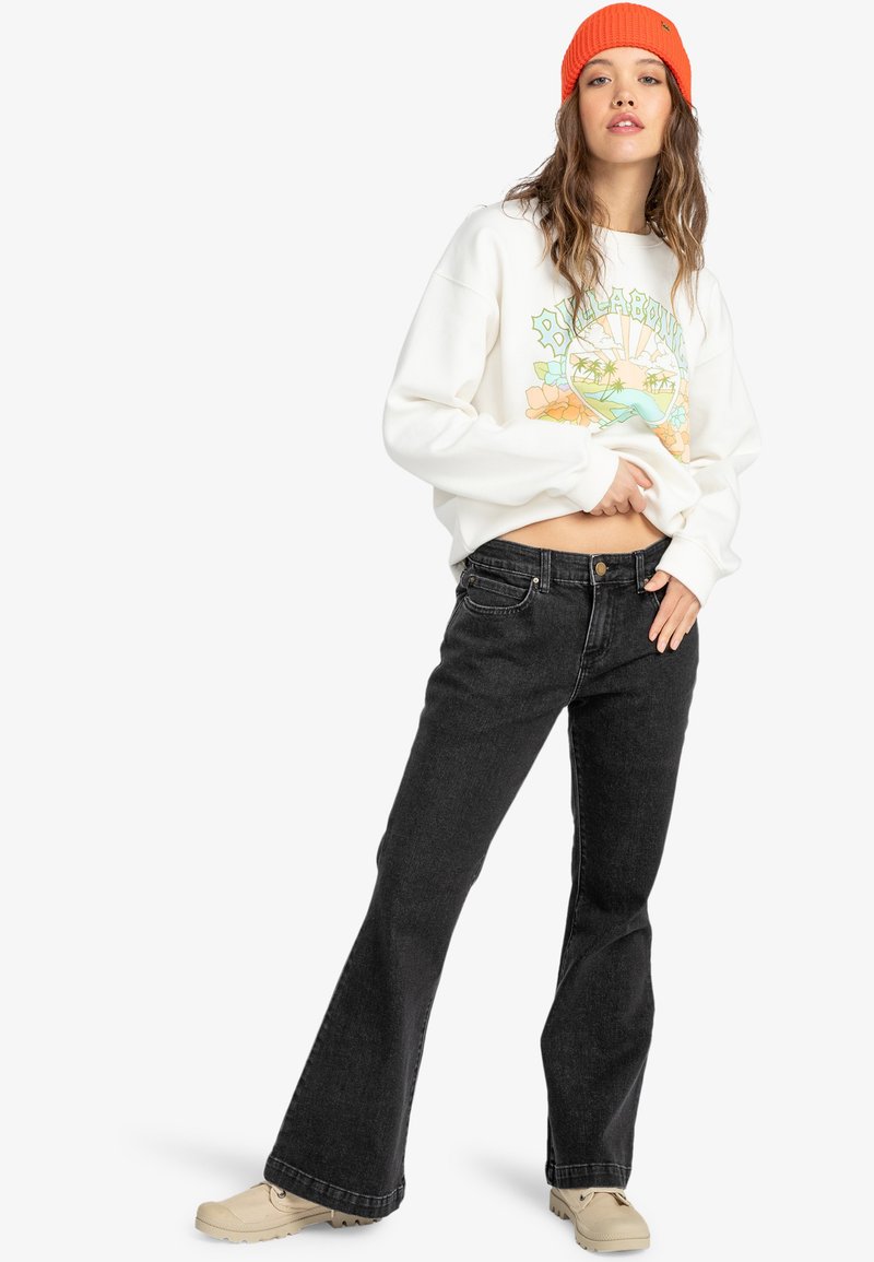 White sweatshirt with colorful graphic design, paired with black flared jeans and beige shoes. Model wears an orange knit beanie.