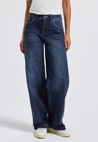 Jean en denim à jambes larges, couleur bleu foncé, avec un design classique à cinq poches, des coutures ton sur ton et une fermeture à bouton à la taille.