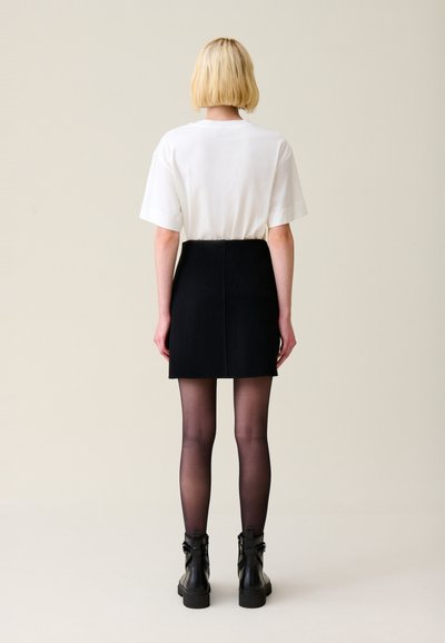 Camiseta blanca de manga corta, combinada con una falda negra de cintura alta. El modelo lleva medias negras transparentes y botas de tobillo.
