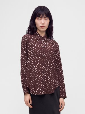 Femme portant une chemise boutonnée marron à manches longues avec des pois rose clair et une jupe noire sur un fond blanc uni.