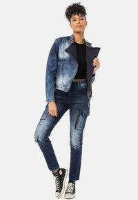 jeansjacke mit einem verblassten blauen Finish, Reißverschluss und gemustertem Kragen, kombiniert mit figurnahen blauen Cargo-Hosen mit Taschen und Reißverschlüssen.