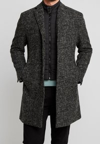 Manteau en laine mélangée texturée grise avec une coupe ajustée, des revers en angle et deux poches avant, superposé sur une doudoune noire.