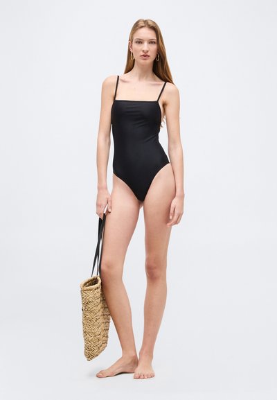 Γυναίκα με μαύρο ολόσωμο μαγιό που κρατάει μια υφασμένη ψάθινη τσάντα με μαύρες χειρολαβές, στέκεται ξυπόλυτη μπροστά σε ένα απλό φόντο.