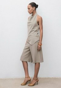 T-shirt sans manches beige avec un col montant, associé à un short de longueur genou de la même couleur. Sandales à talons hauts à lanières marron et un bracelet.