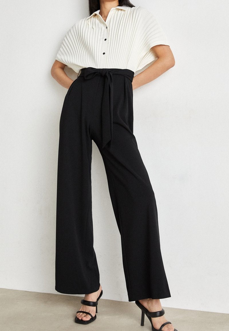 Femme portant un pantalon noir taille haute à jambes larges avec une ceinture nouée, un t-shirt blanc côtelé à manches courtes, et des sandales noires à talons, debout.