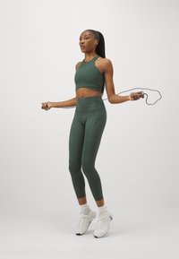 Grønn idrettskort topp og høy midje leggings, laget av elastisk materiale, kombinert med hvite sneakers. Modellen hopper med tau.