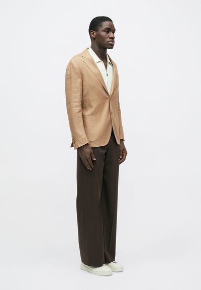 Άνδρας που φοράει μπεζ σακάκι, λευκό πουκάμισο με φερμουάρ, παντελόνι σε σκούρο καφέ με φαρδιά μπατζάκια και λευκά αθλητικά παπούτσια, στέκεται μπροστά σε ένα απλό λευκό φόντο.