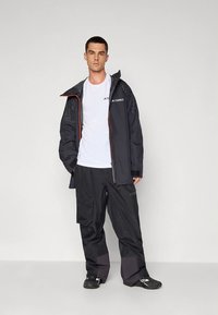 Črna vodoodporna jakna z oranžnimi poudarki, kombinirana s črnimi cargo hlačami z žepi in teksturirano tkanino, nošena čez belo majico.