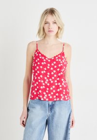 Top rosso senza maniche con motivo floreale bianco, scollatura a onda e spalline sottili, abbinato a jeans a gamba larga blu chiaro.