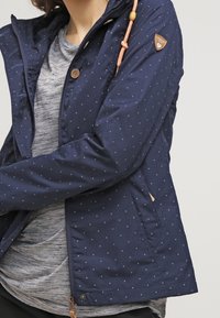 Marinblå jacka med vita prickar, huva, framfickor och beigea togglar, tillverkad av ett lätt och smidigt material.