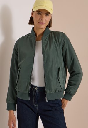 Femme portant une casquette jaune, une veste bomber vert foncé zippée, une chemise blanche et un jean bleu foncé, debout avec une main dans la poche.