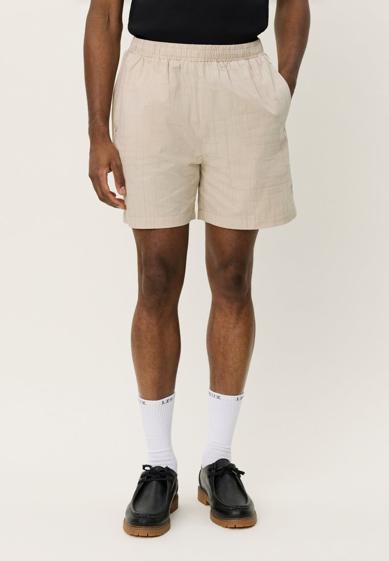 Pantalones cortos beige con una cinturilla elástica, que presentan un sutil patrón de cuadrícula. Combinados con calcetines blancos y zapatos negros para crear contraste.
