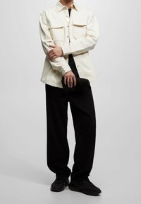 Camisa de botón de crema claro con dos bolsillos en el pecho, combinada con pantalones negros holgados y zapatillas negras, sostenida con un bolso oscuro.