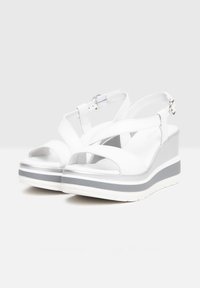 Weiße Keilsandalen mit glänzender Oberfläche, ausgestattet mit überkreuzten Riemen, einer verstellbaren Knöchel- Schnalle und einer grau-weiß gestreiften Plateau-Sohle.