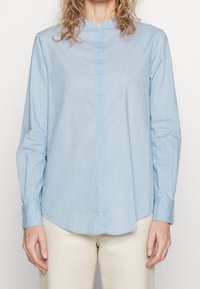 Ljusblå långärmad skjorta i slät tyg, med ståkrage och knappar fram, samt en något rundad fåll.