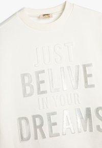 Sudadera blanca con letras texturizadas en relieve. Los acentos de foil plateado dicen "SÓLO CREE EN TUS SUEÑOS". Cuello redondo, puños y dobladillo acanalados.
