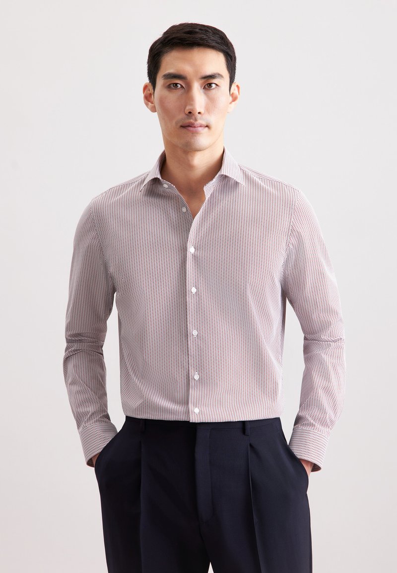 Koszula zapinana na guziki w jasnoróżowym kolorze z drobnym, ciemnoczerwonym wzorem geometrycznym. Posiada kołnierz rozłożysty i długie rękawy, zestawiona z ciemnymi spodniami.