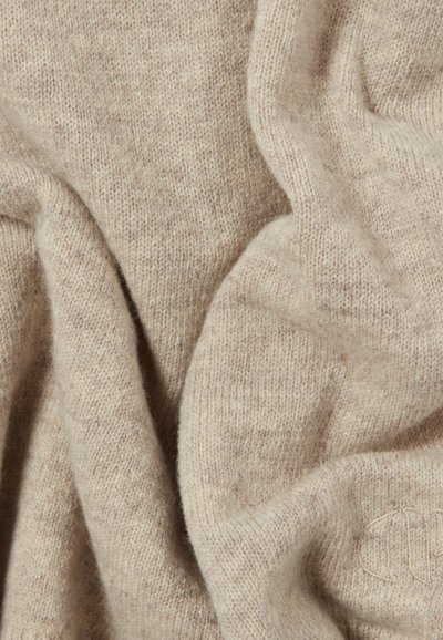 Tessuto di maglia beige con una superficie morbida e testurizzata. Aspetto leggermente melangiato con variazioni sottili nel tono. Presenta un piccolo dettaglio ricamato.