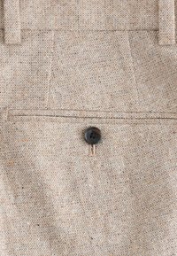 Section de pantalon en tissu beige texturé présentant une poche arrière avec un bouton noir unique et des détails subtilement tachetés.