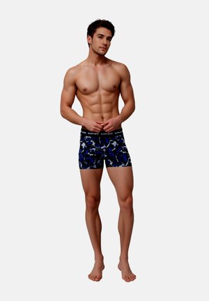 Homme musclé torse nu, debout pieds nus, portant un boxer bleu et noir à motifs sur un fond uni.