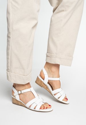 White wedge sandals with crossed straps, cork wedge heel, and an adjustable ankle strap, paired with light beige ribbed pants.