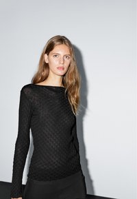 Black long-sleeve top with a textured pattern, boat neckline, and slim fit. Background is neutral with soft lighting.