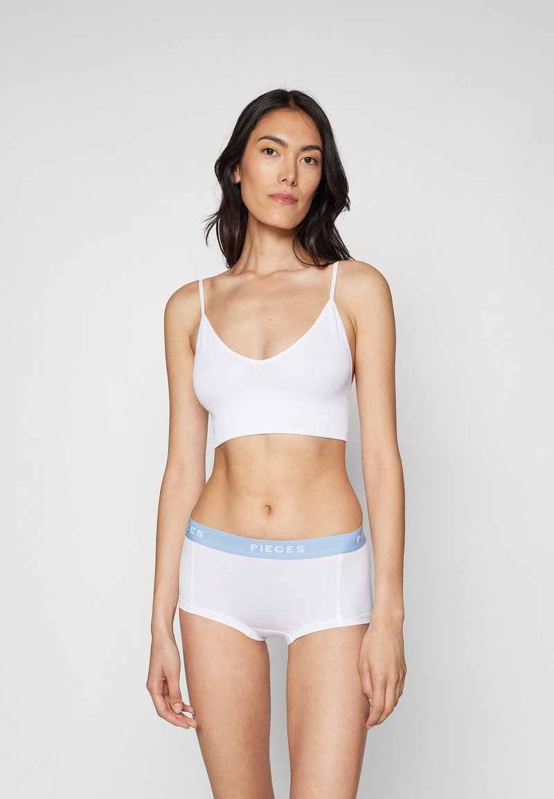 Hvit ribbet crop-top med tynne stropper, kombinert med hvite shorts med en lyseblå midjebånd merket "PIECES." Glatt tekstur.